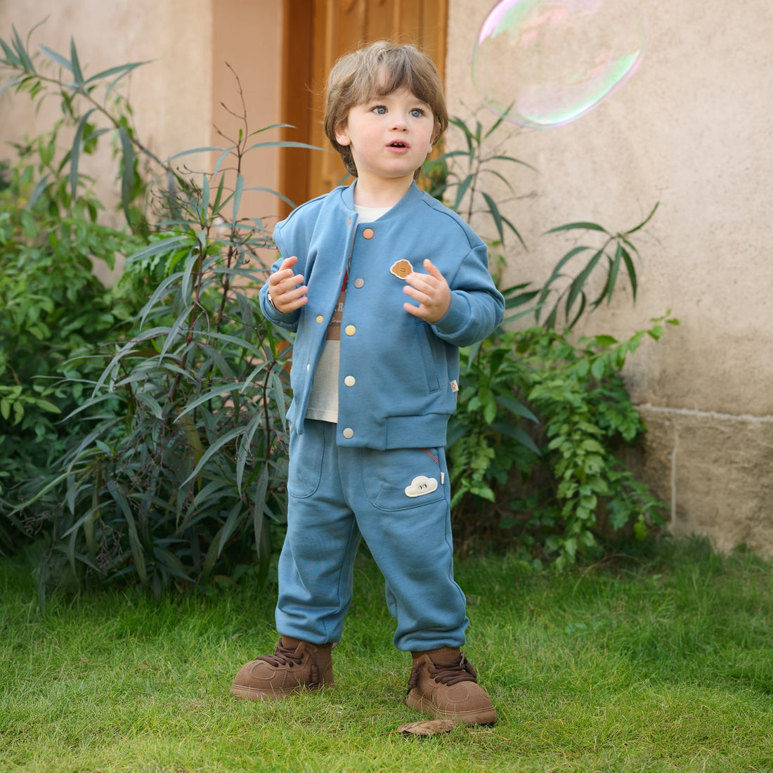 Child wears the Nest Designs Oceanview Smiley Brunch Baseball Jacket with matching blue pants on grass while watching a floating bubble