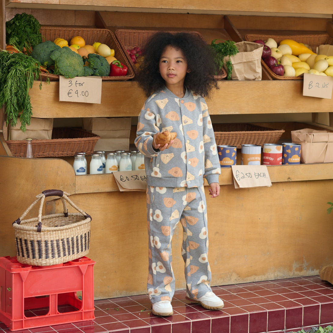 Child models the Nest Designs Smiley Side Up breakfast-print baseball jacket and matching pants at a produce stand beside a woven basket