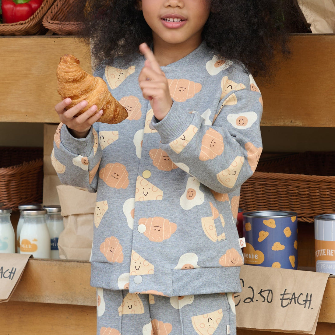 Nest Designs Smiley Side Up baseball jacket close-up as a child holds a croissant, showing the grey allover breakfast-character print