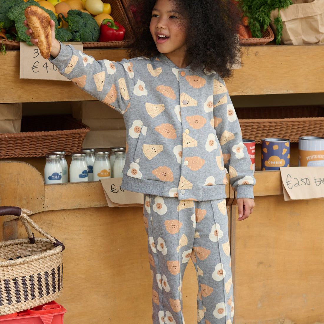 Child reaches toward produce in the Nest Designs Smiley Side Up breakfast-print baseball jacket and matching pants at a market stall