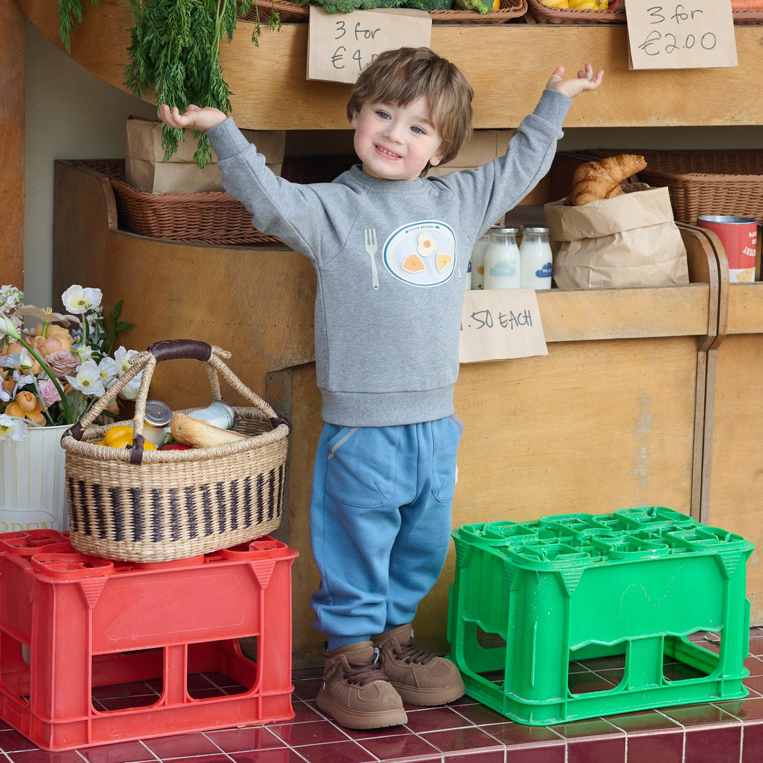 Nest Designs Smiley Plate Velcro Sweatshirt in Pebble Grey, worn by a child raising both arms between market crates