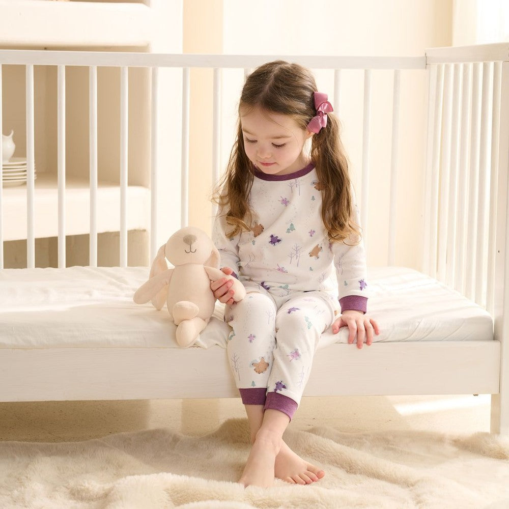 Nest Designs Snow Angels two-piece long-sleeve pajama set; white with purple cuffs, girl sits on bed with plush bunny