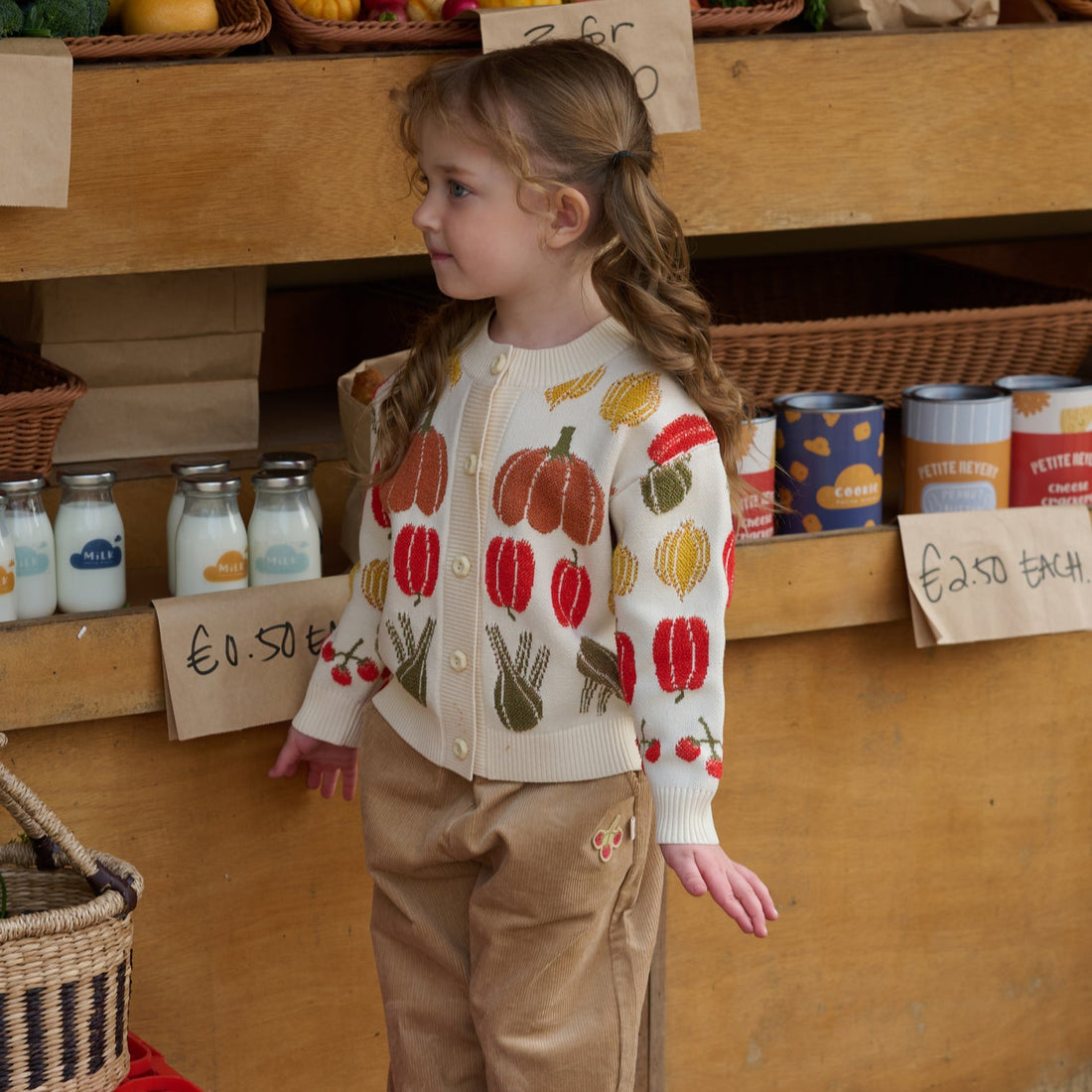 Nest Designs Veggie Jacquard Cardigan in Veggie Tales cream worn in profile with tan trousers at a produce market