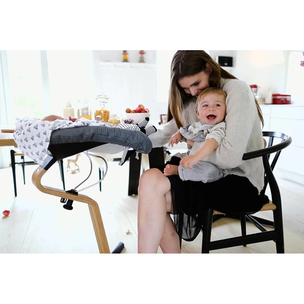 NOMI Chair White Oak high chair in a bright kitchen; mother with a smiling baby nearby shows its compact, modern wood design.