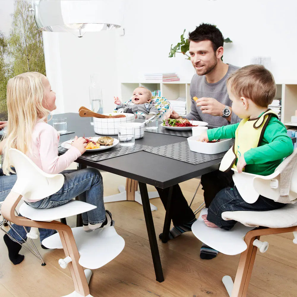 NOMI Highchair White Oak at a bright family mealtime, where a dad feeds the baby and siblings eat.
