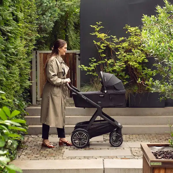 Nuna DEMI next Bassinet — Woman pushing Nuna DEMI next stroller with Caviar bassinet on a garden walkway