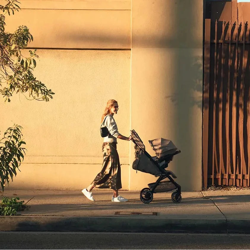 Nuna TRVL LX Stroller + PIPA Urbn Infant Car Seat Travel System showing Woman walking with a stroller against a beige building with the number 1443.
