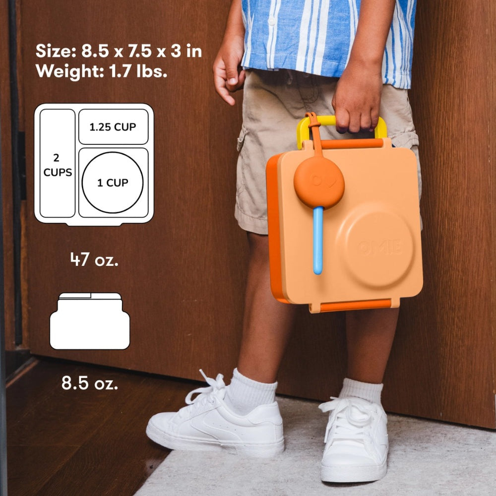 OmieBox Version 2 lunch container in orange/beige carried by a kid with a blue utensil, showing its 2-cup/1-cup compartments.