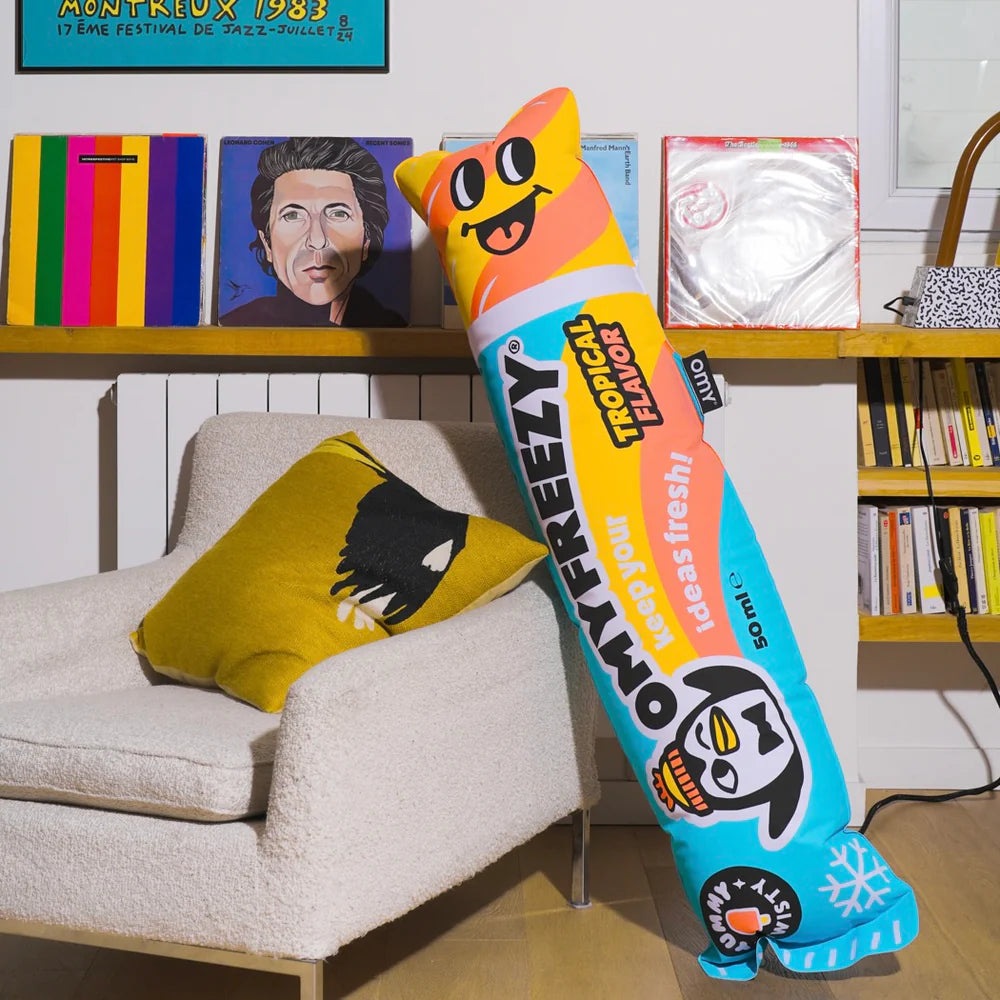 OMY Freezy Giant inflatable pillow, blue/orange with cartoon penguin, leaning against a cream sofa.