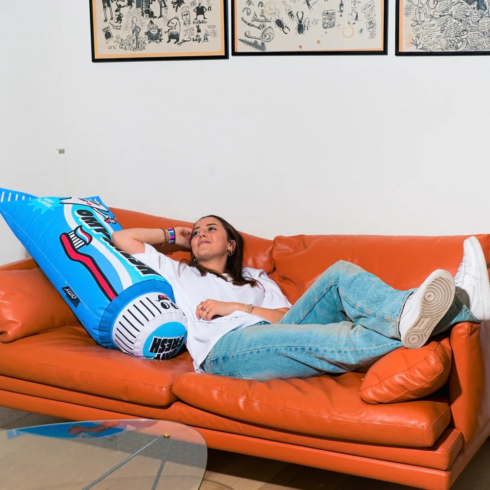 OMY Fresh Giant inflatable pillow on a bright orange sofa; woman lounges, showing its oversized blue cushion for relaxation.
