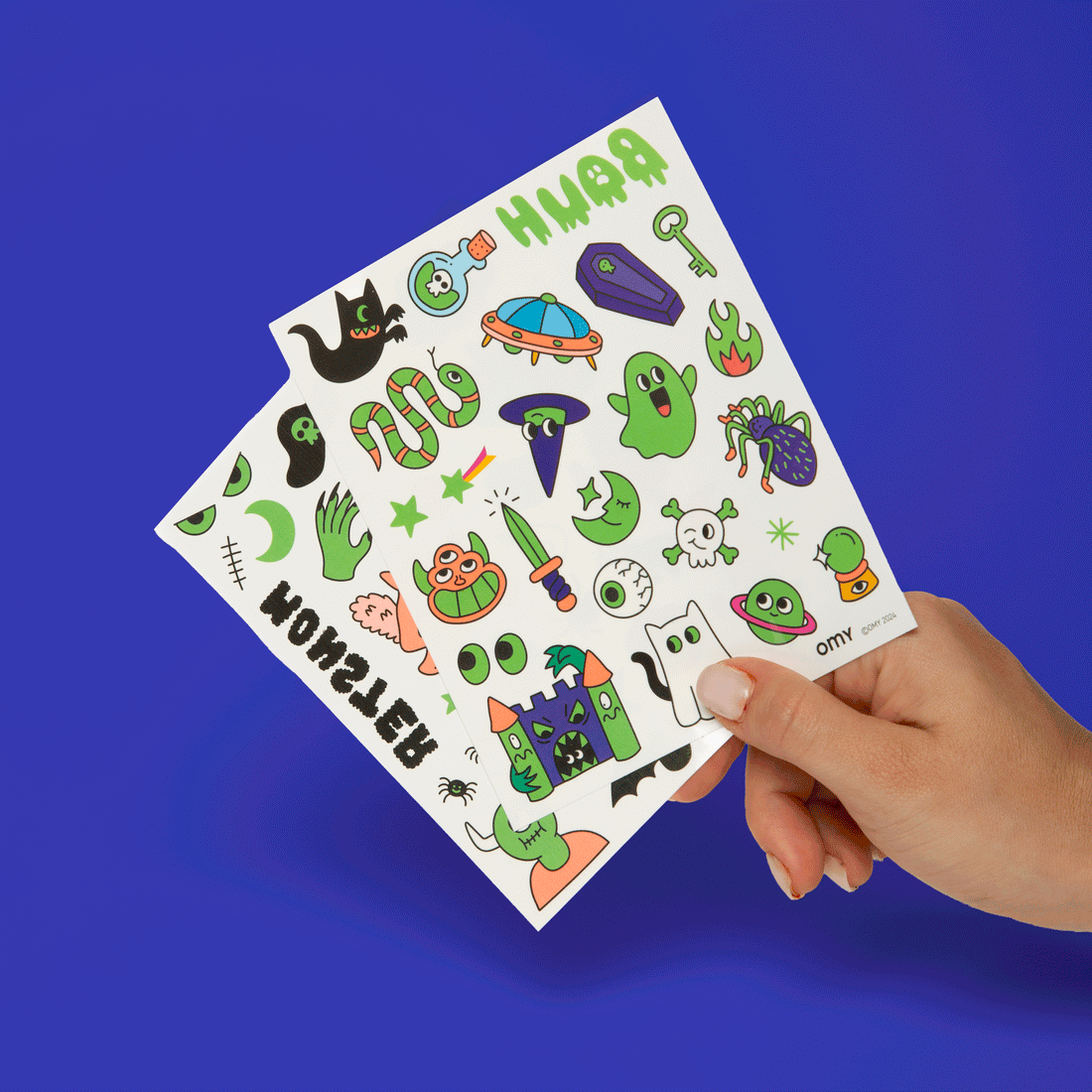 OMY Glow in the Dark Tattoos sheet held by hand, showing bright playful cartoon designs.