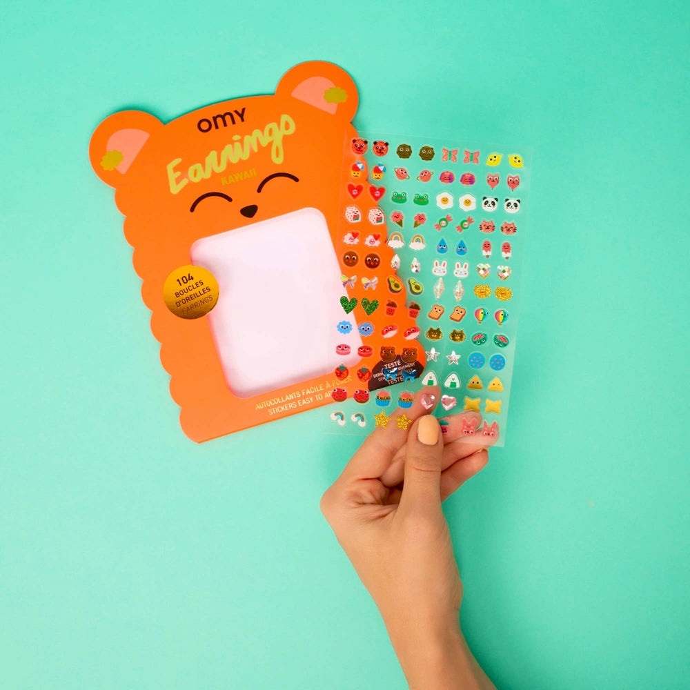 OMY Kids Sticker Earrings - Kawaii in bright orange bear packaging; a hand holds vibrant sticker sheets ready to wear.