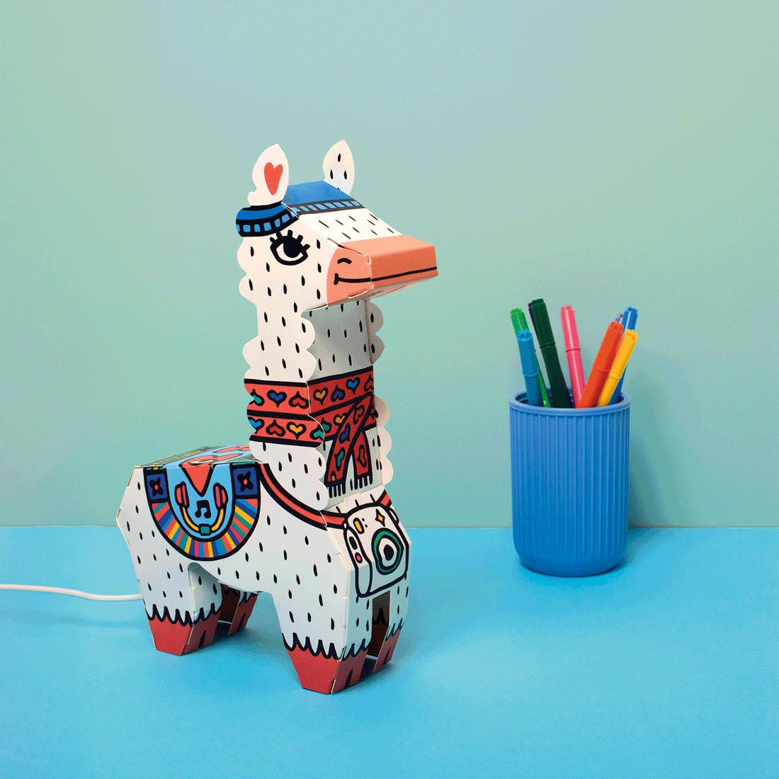 OMY - Lama - 3D Lamp: white dotted llama with colorful accents on a pastel blue desk beside a blue cup of markers.