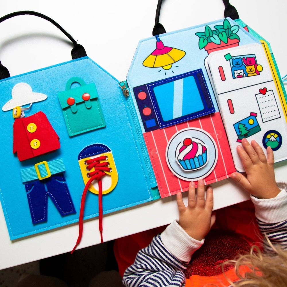 OMY Montessori Play Kit, vibrant fabric activity book; child turns a colorful page with fridge magnets.