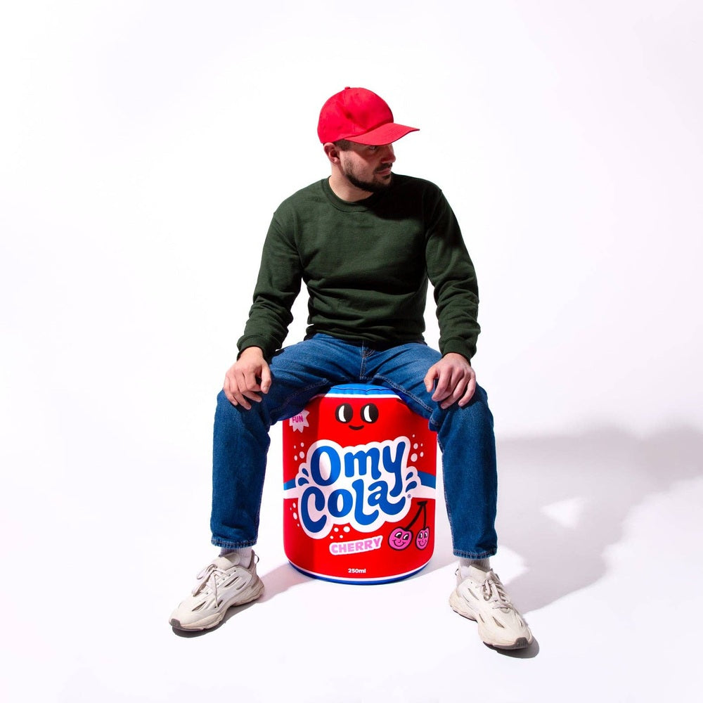 OMY Cola Giant inflatable pillow; red can with blue logo, man in green sweater and red cap seated on it.