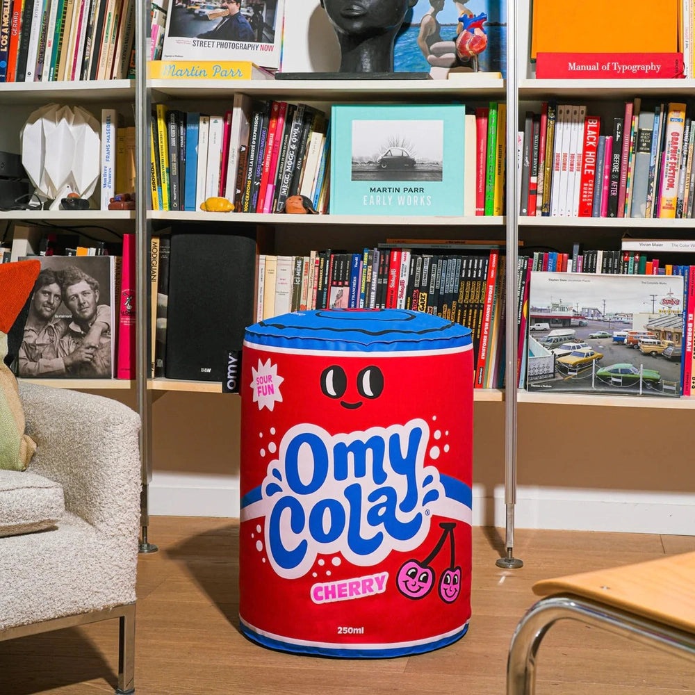 OMY Cola Cherry inflatable pillow in red can design with blue top used as a playful floor pouf in a book-filled living room.