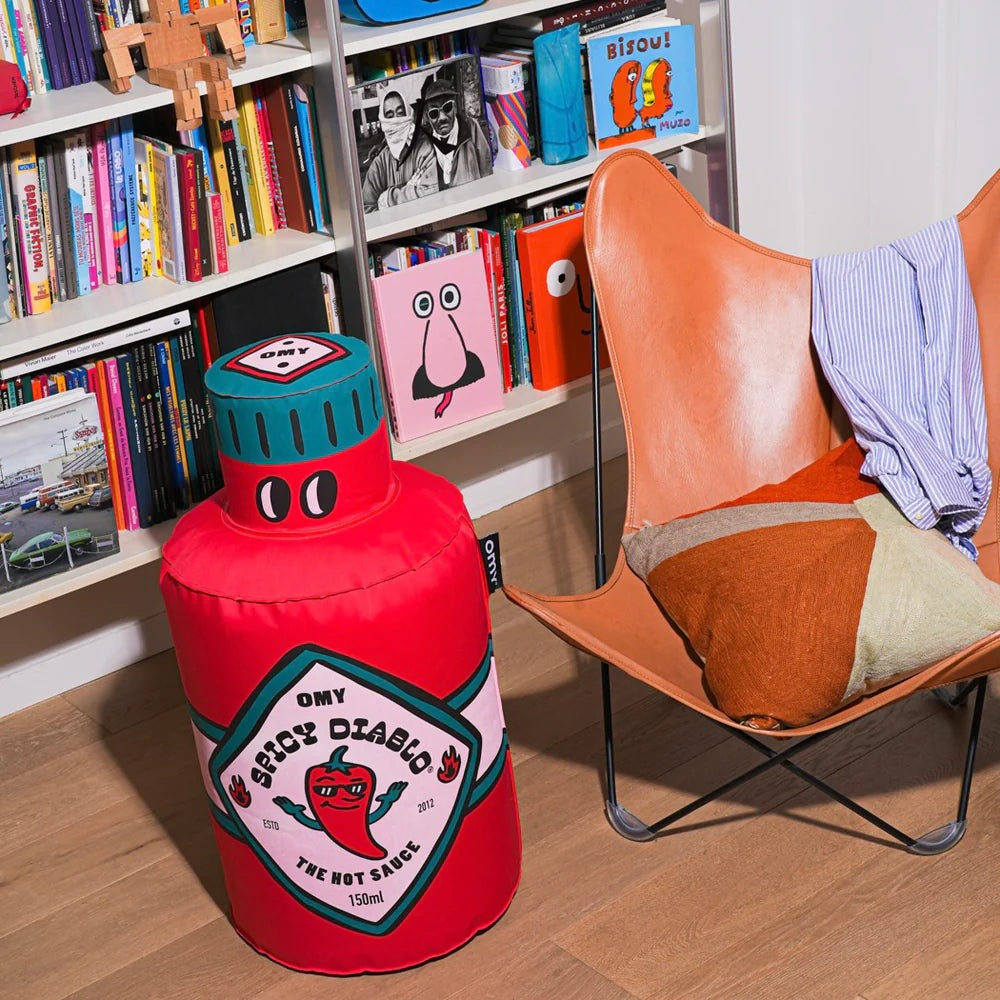 OMY Spicy Diablo Giant Inflatable Pillow, red with teal cap, propped beside an orange chair in a cozy reading nook.