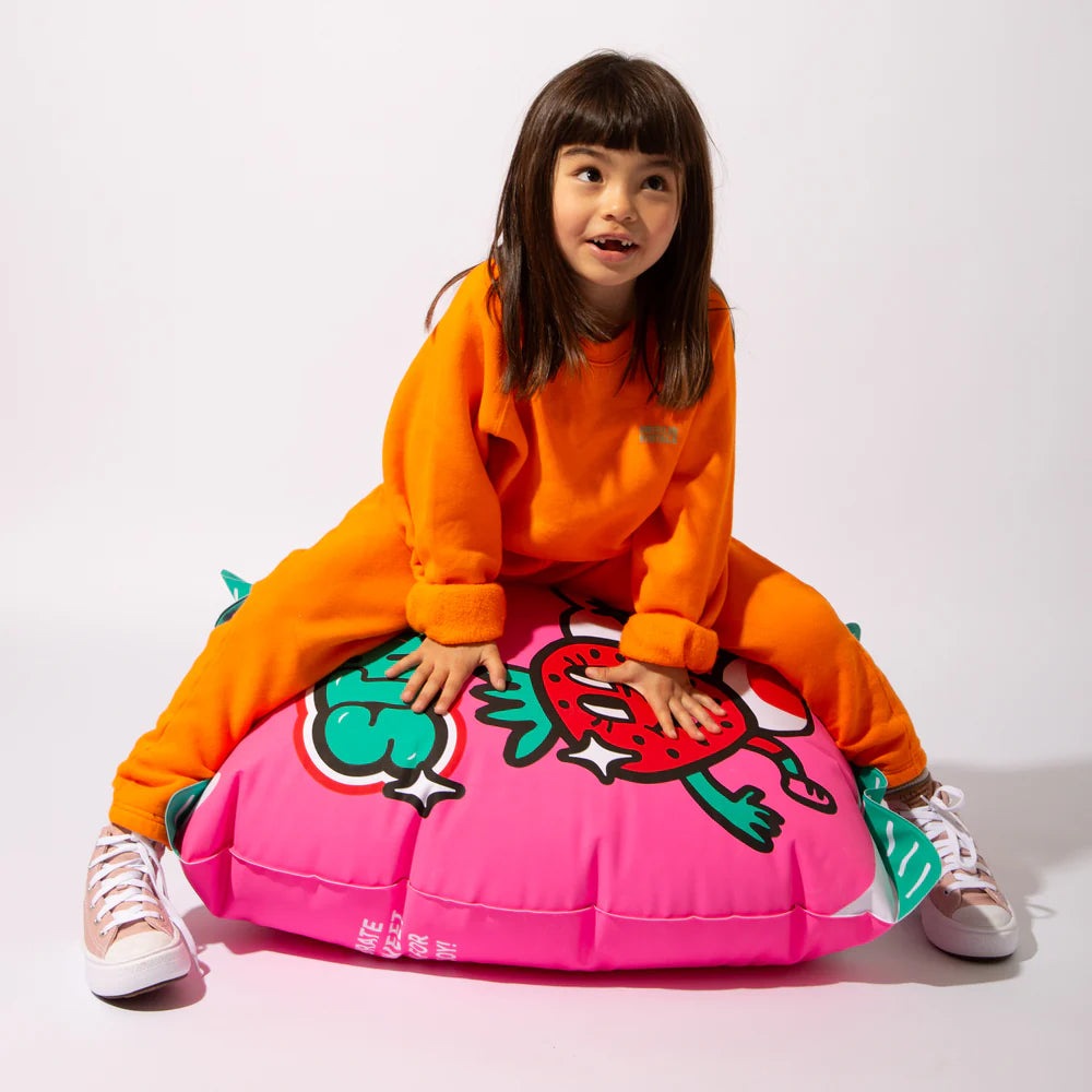 OMY Strawbies Giant inflatable pillow in pink with cartoon strawberry, as a child in orange sits atop it.