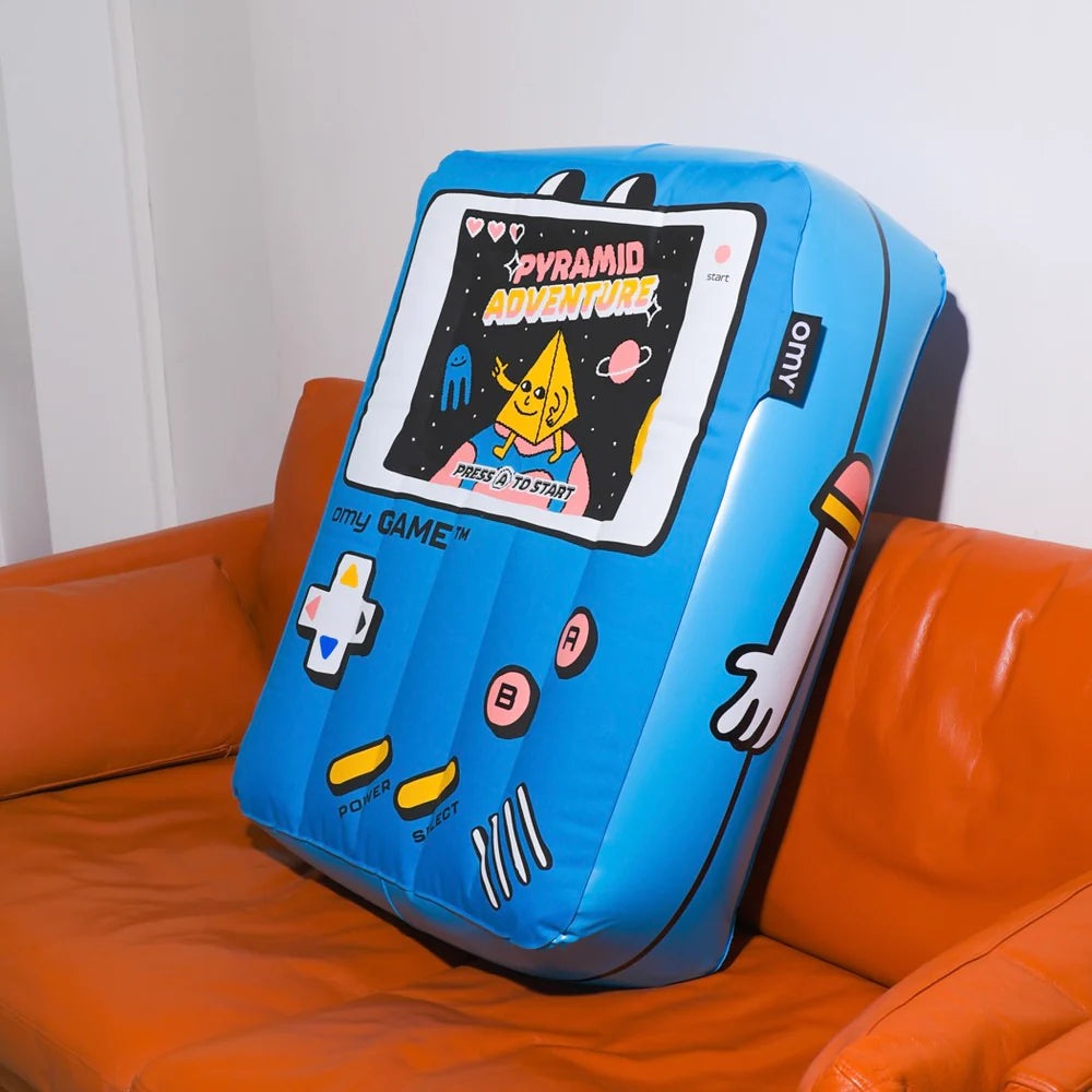 OMY Giant inflatable pillow in blue, retro handheld game design with screen and controls, resting on an orange sofa.