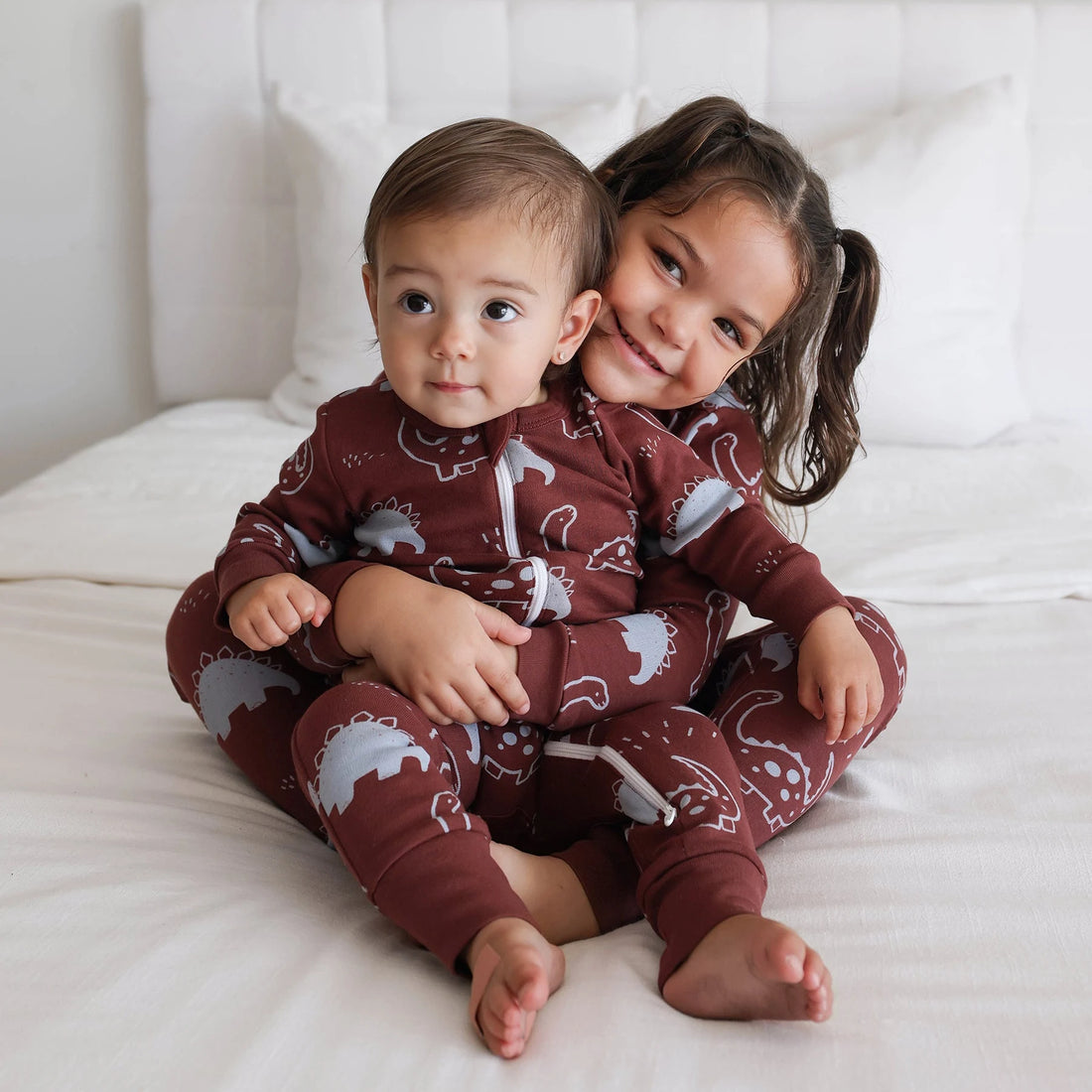 Two children wearing Parade Chocolate Dinosaurs organic two-way zip rompers on white bedding.