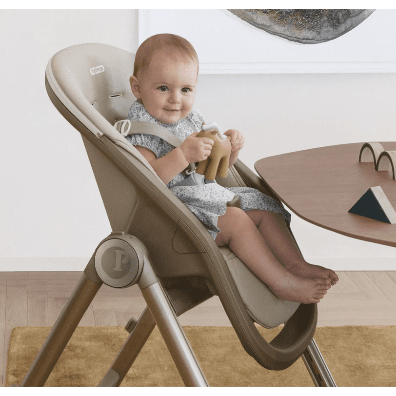 Peg Perego Living High Chair in beige, baby seated with safety harness, gripping a toy near a wooden table.