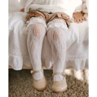 Petite Maison Kids Lola Lace Socks on a toddler, white knee-high lace socks, sitting with feet dangling.