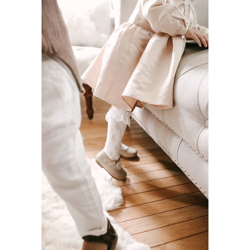 Petite Maison Kids Lola Lace Socks knee-highs in white lace with scalloped trim, shown as a child lifting her foot to reveal the pattern.