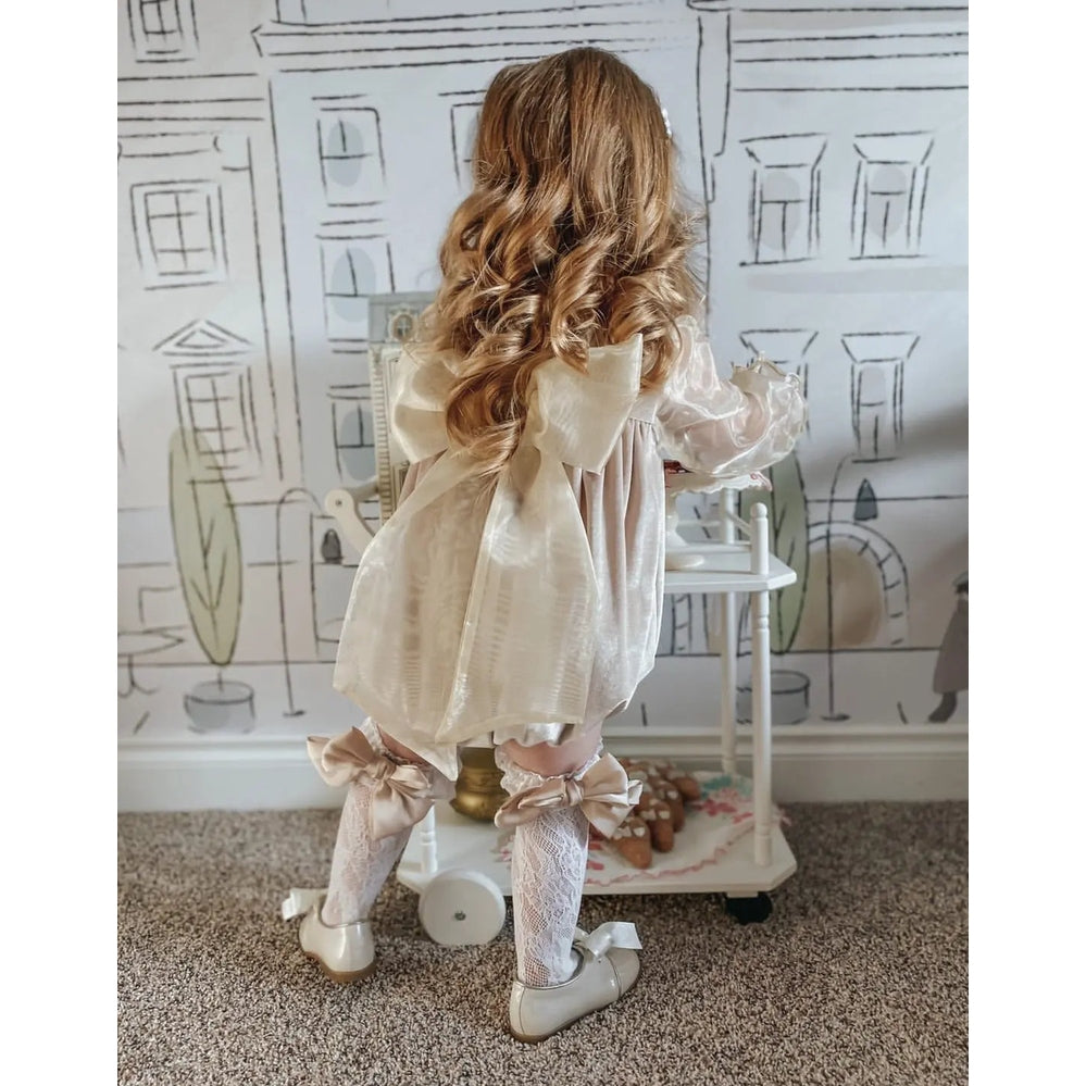 Petite Maison Kids Lola Lace Socks With Satin Bows in cream, worn by a girl standing in a pastel room with lace socks tied in satin bows.