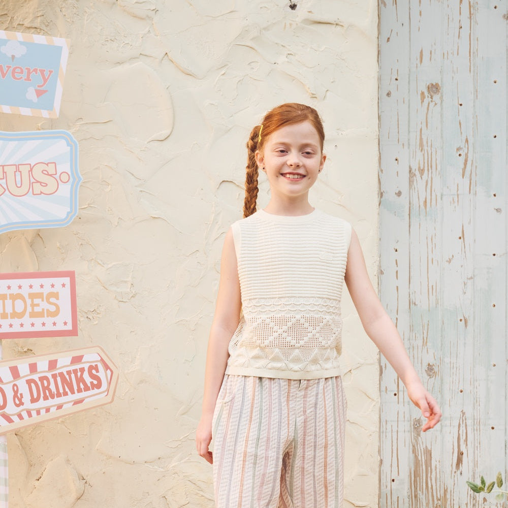 Young girl in a sleeveless top and striped pants standing outdoors with rustic background