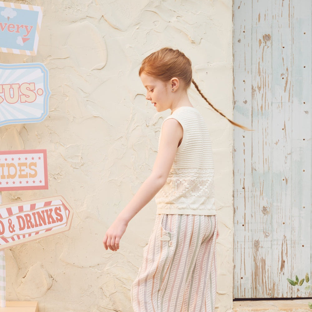 Young girl in a sleeveless top and striped pants standing outdoors near a rustic wooden wall with decorative signs.