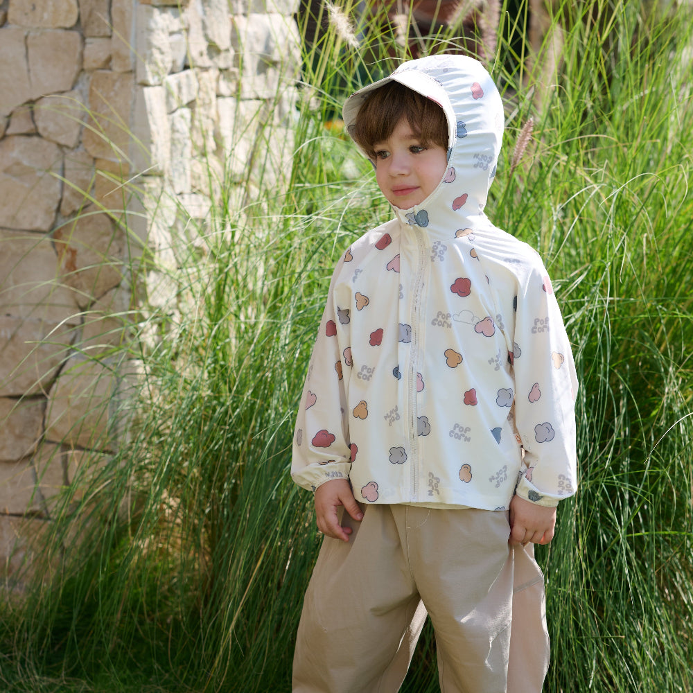 Child wearing a patterned raincoat standing outdoors near a stone wall and tall grass.