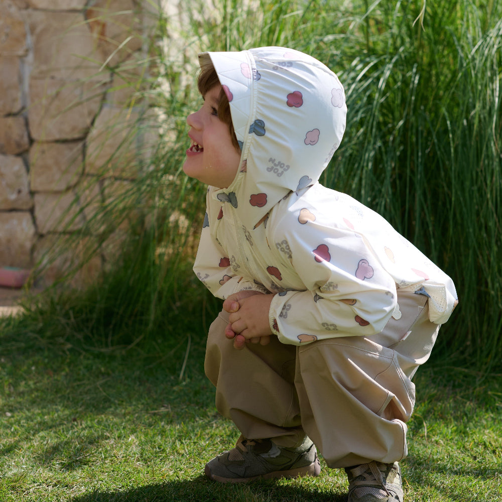 Child in a raincoat and bonnet outdoors near a stone wall and grass.