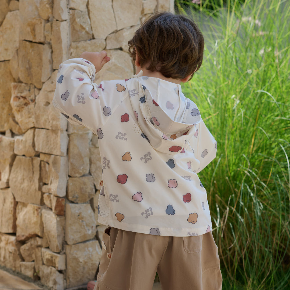 Child wearing a patterned shirt and beige pants standing near a stone wall and grass.