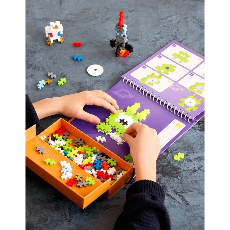 Plus-Plus Activity Pad Space 125pcs; child builds a green pixel figure on purple guidepad with orange storage box.
