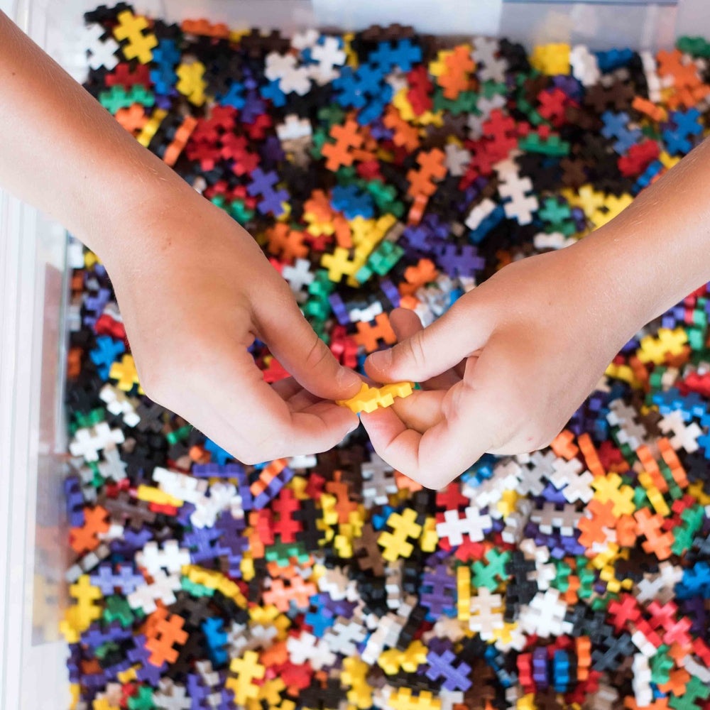 PLUS-PLUS Mini 3600 Educational construction toy; hands connect a yellow-orange piece to colorful interlocking bricks.