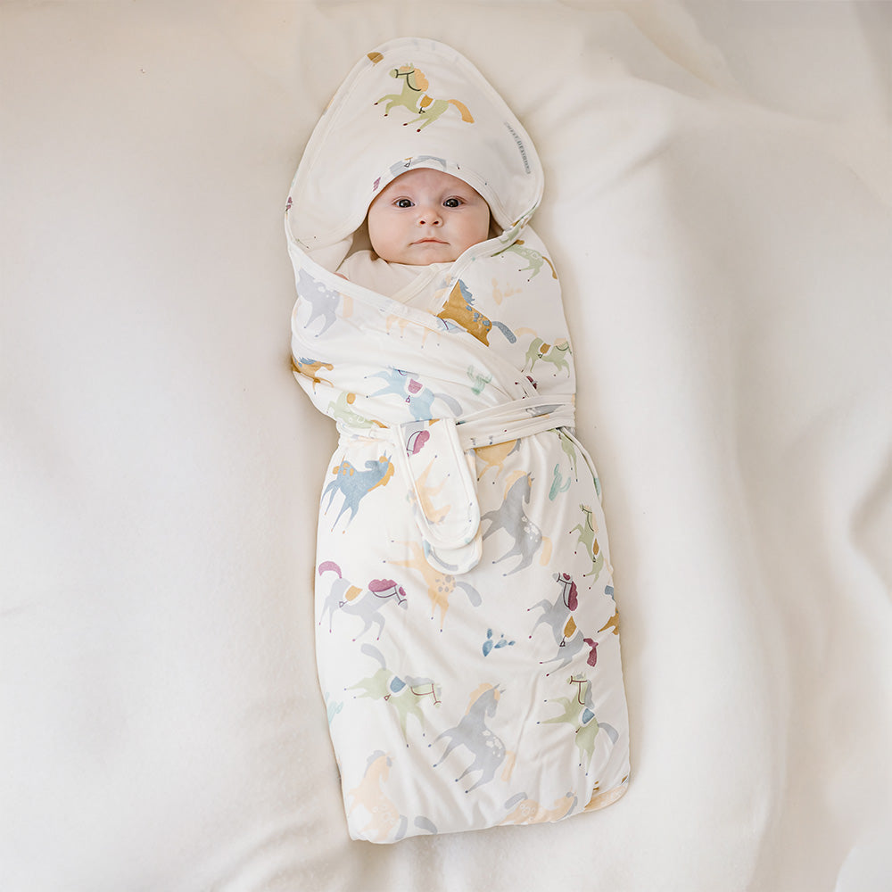 Nest Designs Pony Express Collector's Bundle Swaddle in Cream with pastel pony print, baby wrapped in hood.