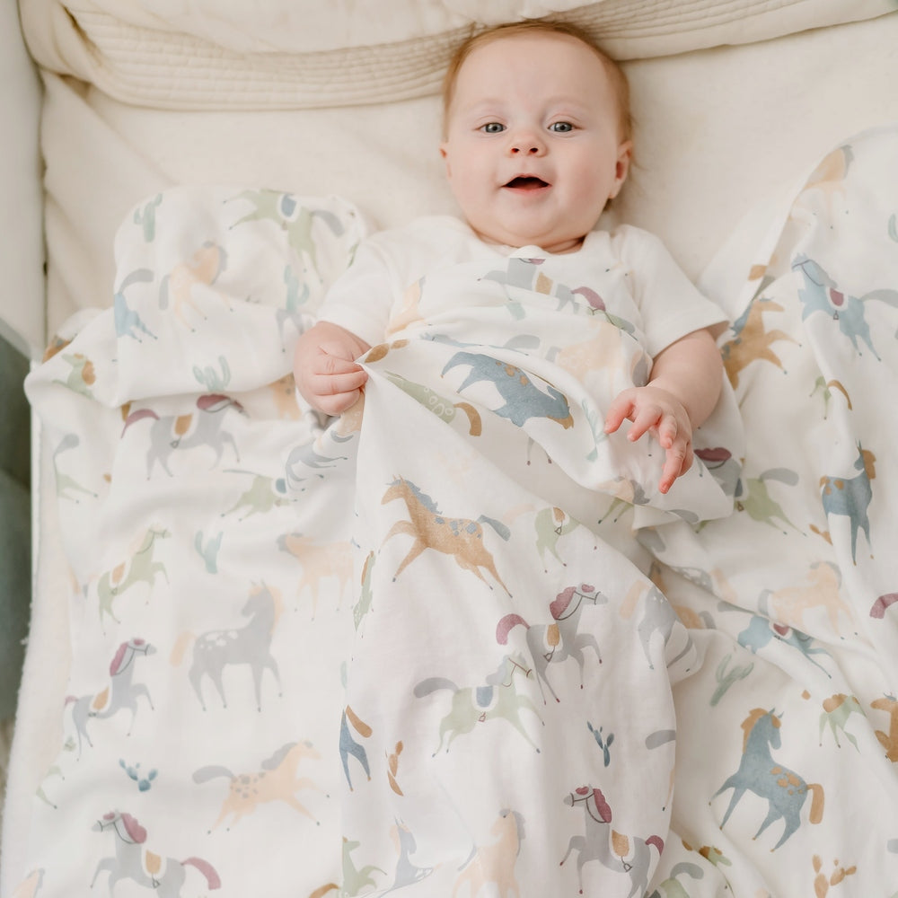 Nest Designs Pony Express Collector's Bundle in White with pastel horse print blanket wrapped around baby.