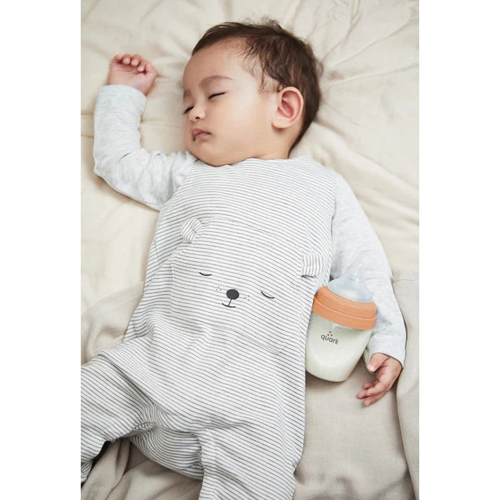 Quark Buubibottle Max Hybrid feeding bottle, 1 pack, beside a sleeping baby in a striped onesie with a peach-cap bottle
