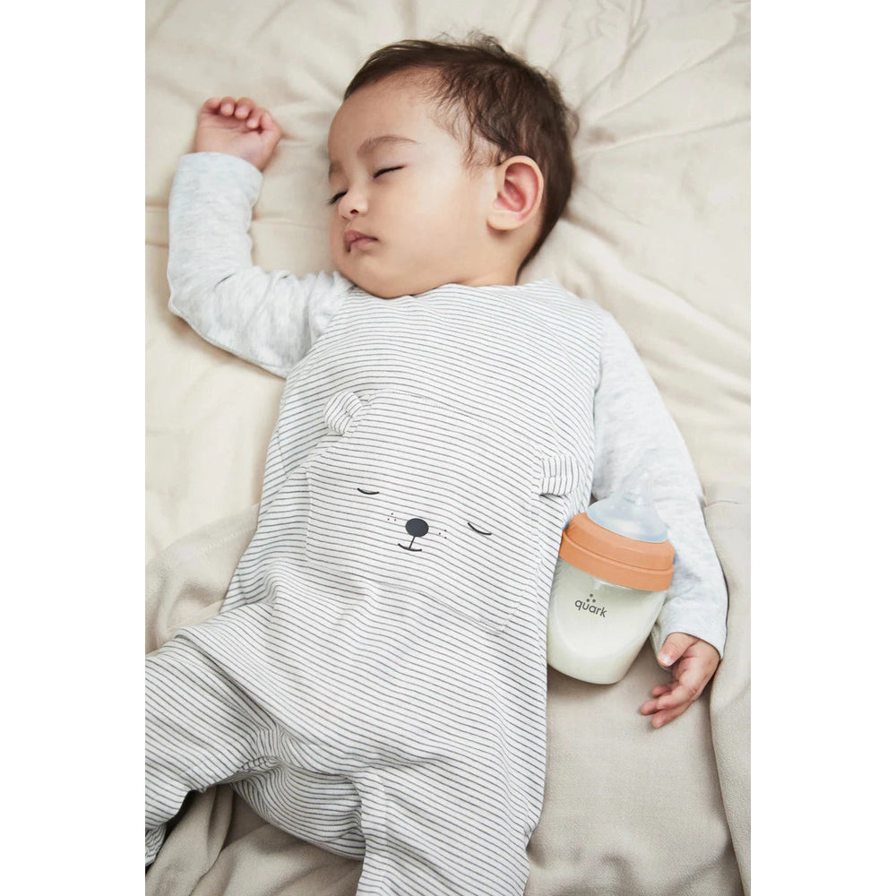 Quark Buubibottle Max Hybrid Feeding Bottle 2 Pack beside a sleeping baby in a striped onesie, orange-cap bottle shown.