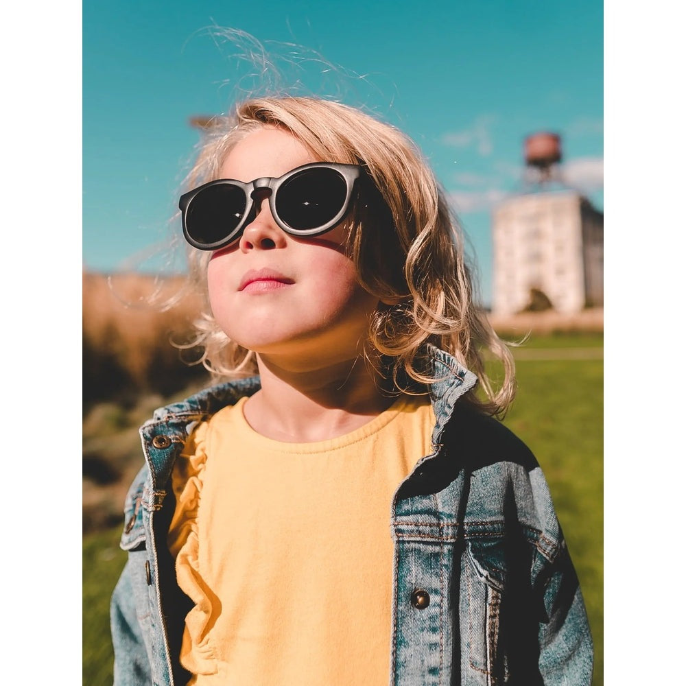 Real Shades Chill Sunglasses Kids Black worn by a child outdoors in a denim jacket.