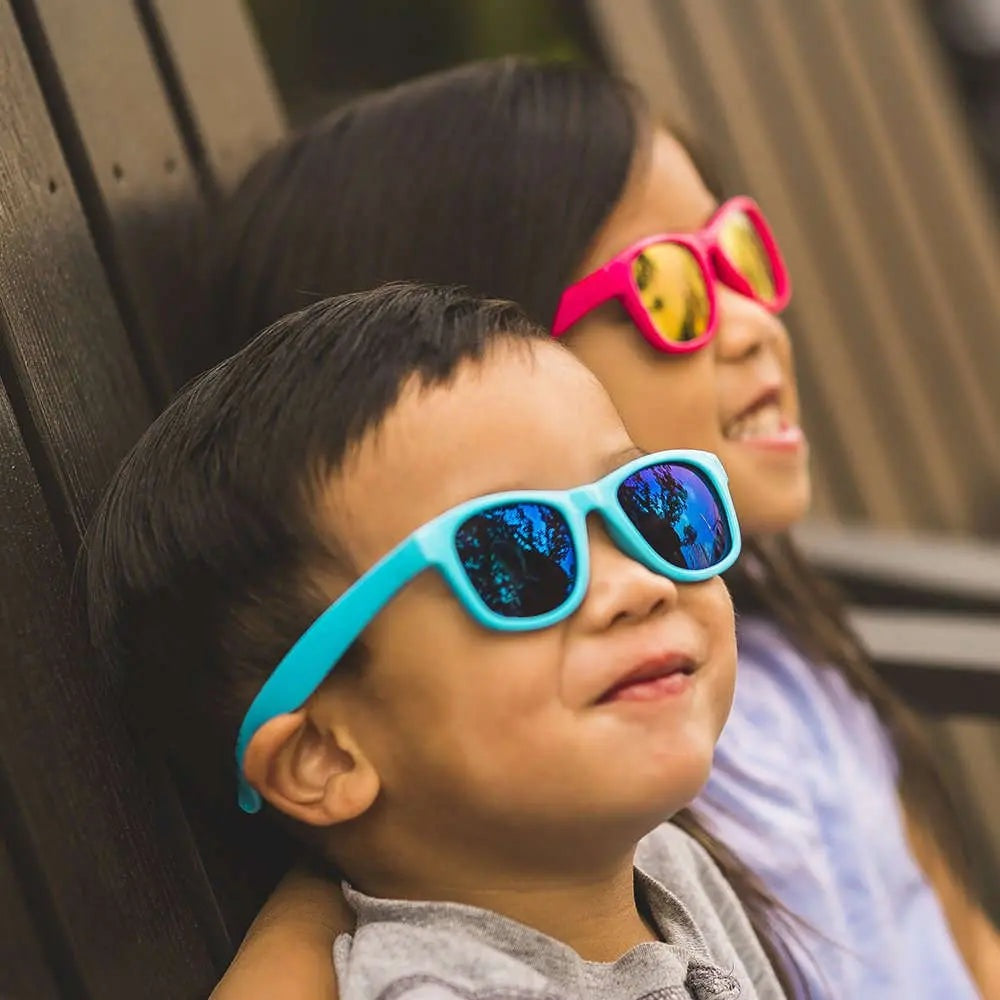 Real Shades Surf Sunglasses Kids Neon Blue with reflective lenses; two kids smile while leaning on a wooden wall in neon blue and pink frames.