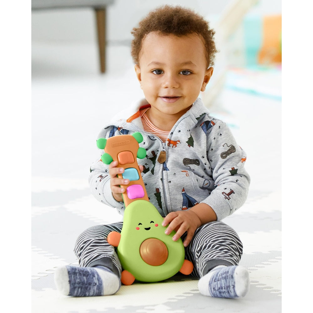 Skip Hop Farmstand Rock A Mole Guitar toy in green avocado with colorful buttons, held by a toddler