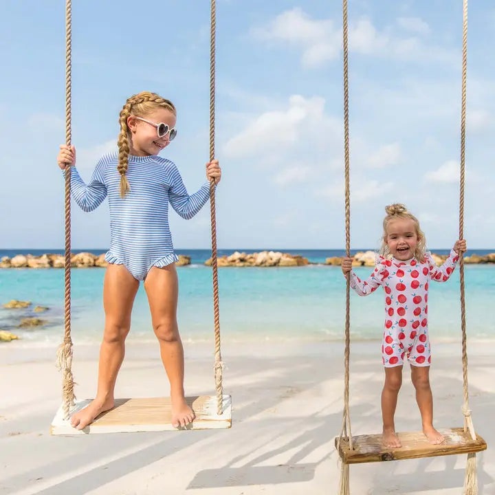 Snapper Rock Cornflower Stripe Surf Suit; two kids in blue-striped and cherry-print swimwear swing on the beach.