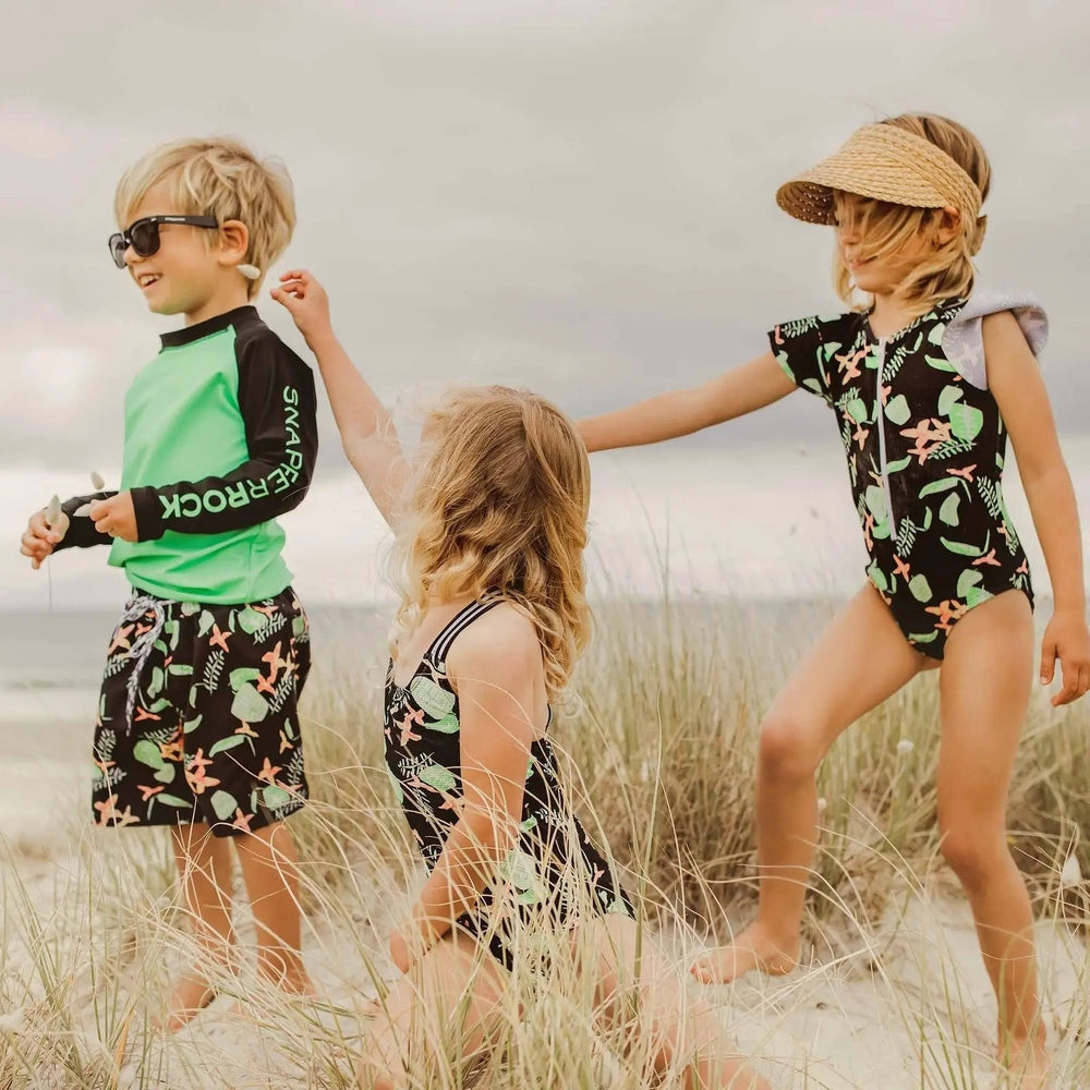 Snapper Rock Neon Rainforest Volley board shorts, black with neon-green rainforest print, worn by kids playing on the beach.