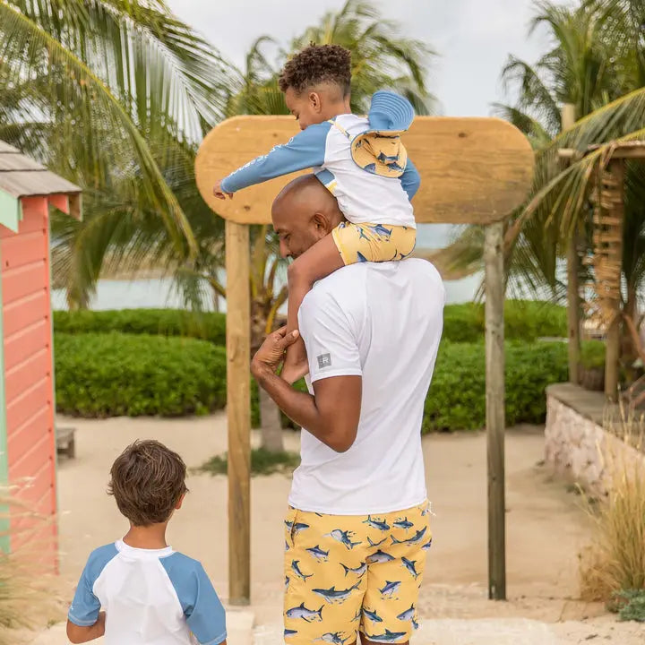 Snapper Rock Sunrise Shark Baby Set: yellow shark-print swim shorts with white/blue top, worn by a toddler perched on dad's shoulders at the beach.