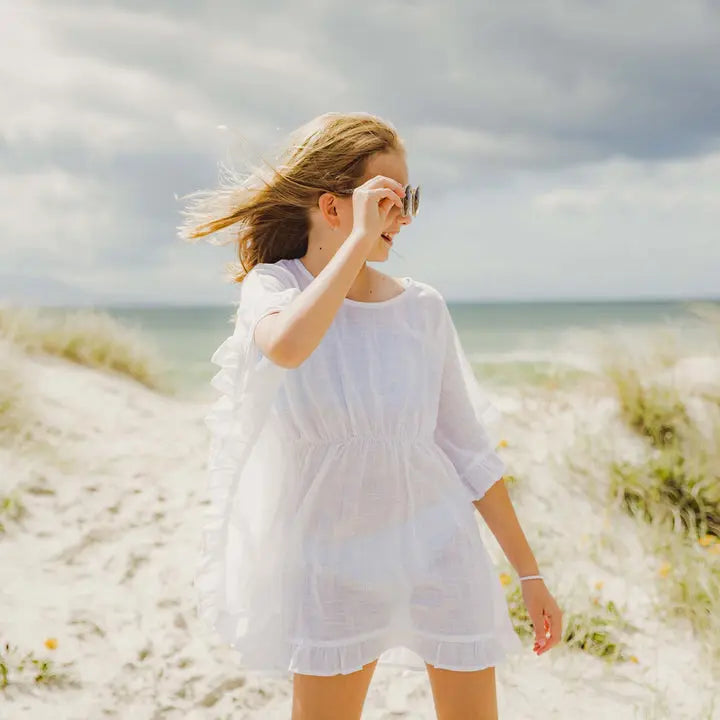 Snapper Rock White Frilled Cover Up: a woman strolls along a windy beach, adjusting sunglasses in the airy white cover-up.