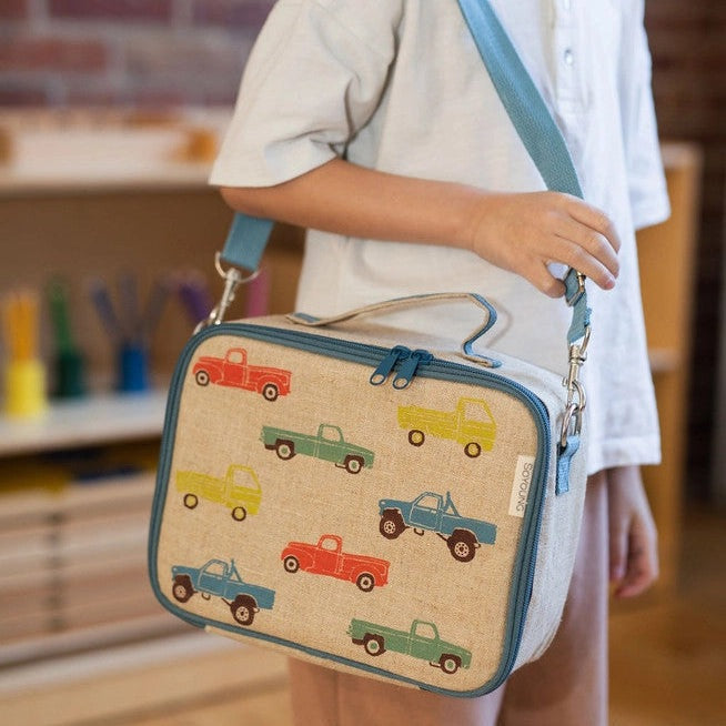 So Young Lunch Box in beige canvas with blue trim and colorful truck pattern, carried by a child using the blue shoulder strap.