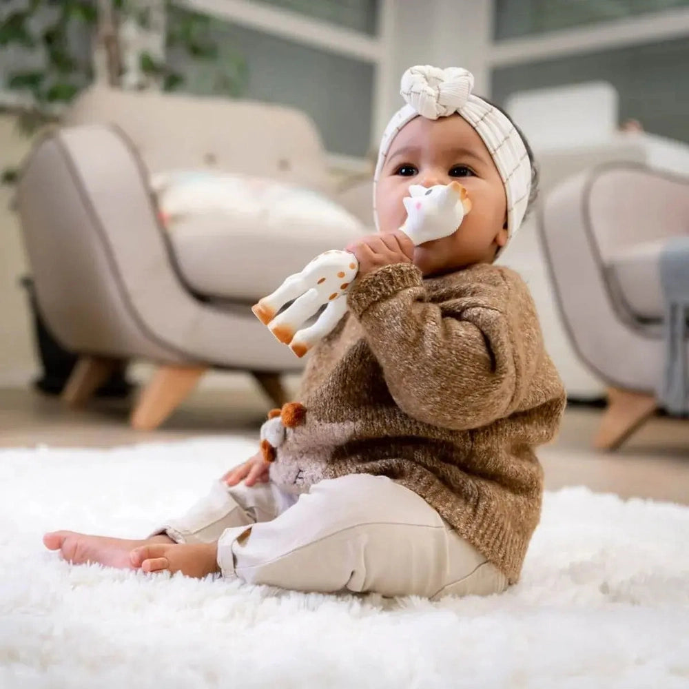 Sophie La Girafe teether held by a baby in a brown knit sweater; white giraffe with orange spots