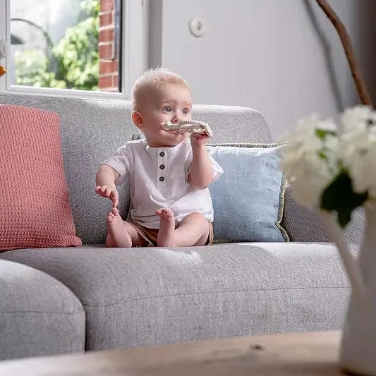 Sophie la Girafe Teething Ring, wooden baby teether, held by a baby on a gray sofa.