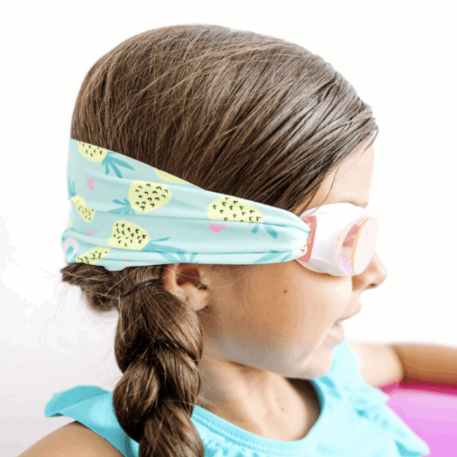 Splash Swim Goggles Pineapple Crush with pink-framed lenses on a girl wearing a turquoise pineapple-print headband.